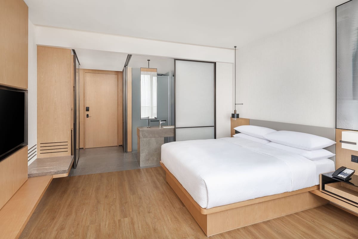 A modern hotel room with a wooden floor, a large bed with white linens, a bedside table with a lamp, and a TV.
