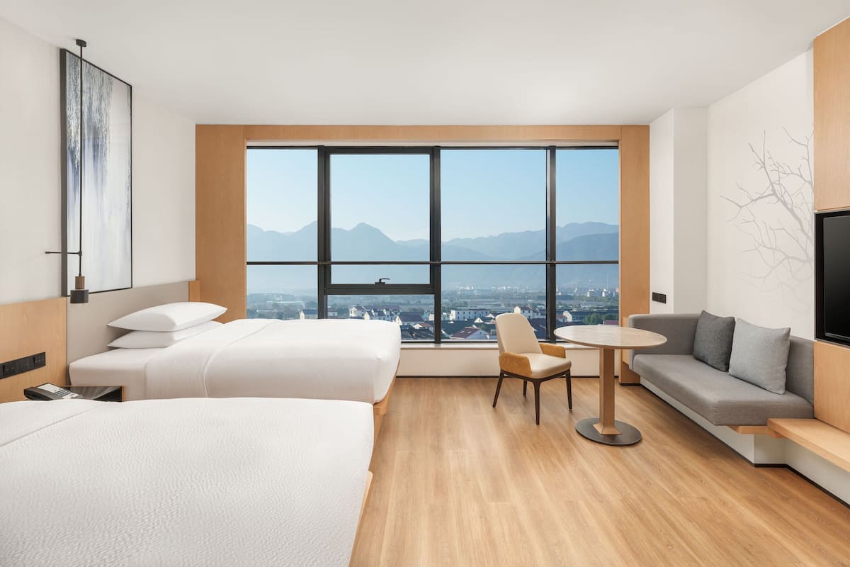 A hotel room with two beds, a sofa, a TV, and a large window offering a city view.