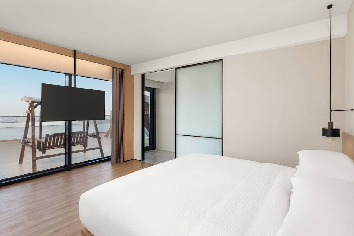 A modern hotel room with a large bed, a TV on a stand, and a view of the city through large windows.