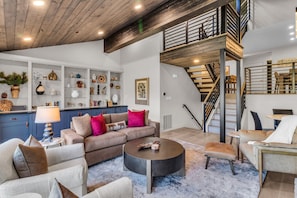 Un vaste espace de vie comprenant un ensemble de canapés, une table basse et une bibliothèque remplie d’objets décoratifs. Un escalier doté d’une rampe en métal mène à l’étage supérieur.