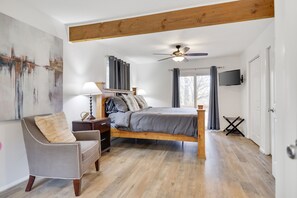 A bedroom with a wooden bed, a chair, a nightstand, a ceiling fan, a TV, and a large abstract painting. - Poconos Lakeside:  Hot Tub & Game Room! Steps Away from Lake Beach and Pool (Albrightsville)