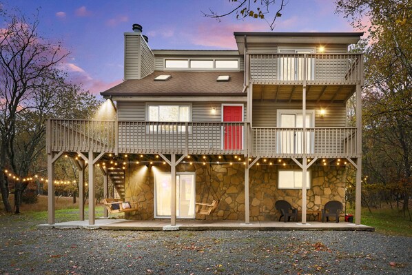 A two-story house with a stone facade, a red door, and a balcony with string lights. - Poconos Lakeside:  Hot Tub & Game Room! Steps Away from Lake Beach and Pool (Albrightsville)
