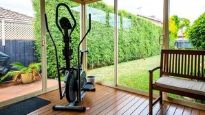 Un patio cubierto con una bicicleta estática, un banco y vistas a un jardín cuidado.