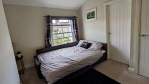 Quarto com cama, janela com cortinas, porta e uma pequena planta em uma mesinha de cabeceira.