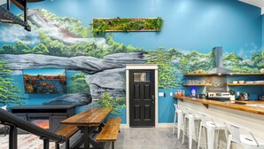 A restaurant with a mural of a waterfall and forest, a black door, a wooden table with benches, and white bar stools.