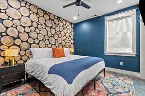 Een slaapkamer met een bed, een nachtkastje, een lamp, een plafondventilator, een raam met zonwering, en een muur bekleed met houten balken.