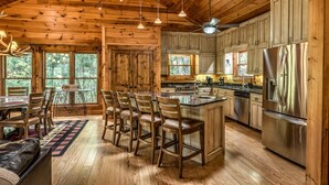 A cabin interior with a wooden dining area, bar stools, a kitchen with stainless steel appliances, and large windows offering a view of the forest.