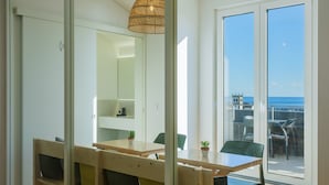 Een moderne eetruimte met een houten tafel en banken, een hanglamp en uitzicht op zee door grote glazen deuren.