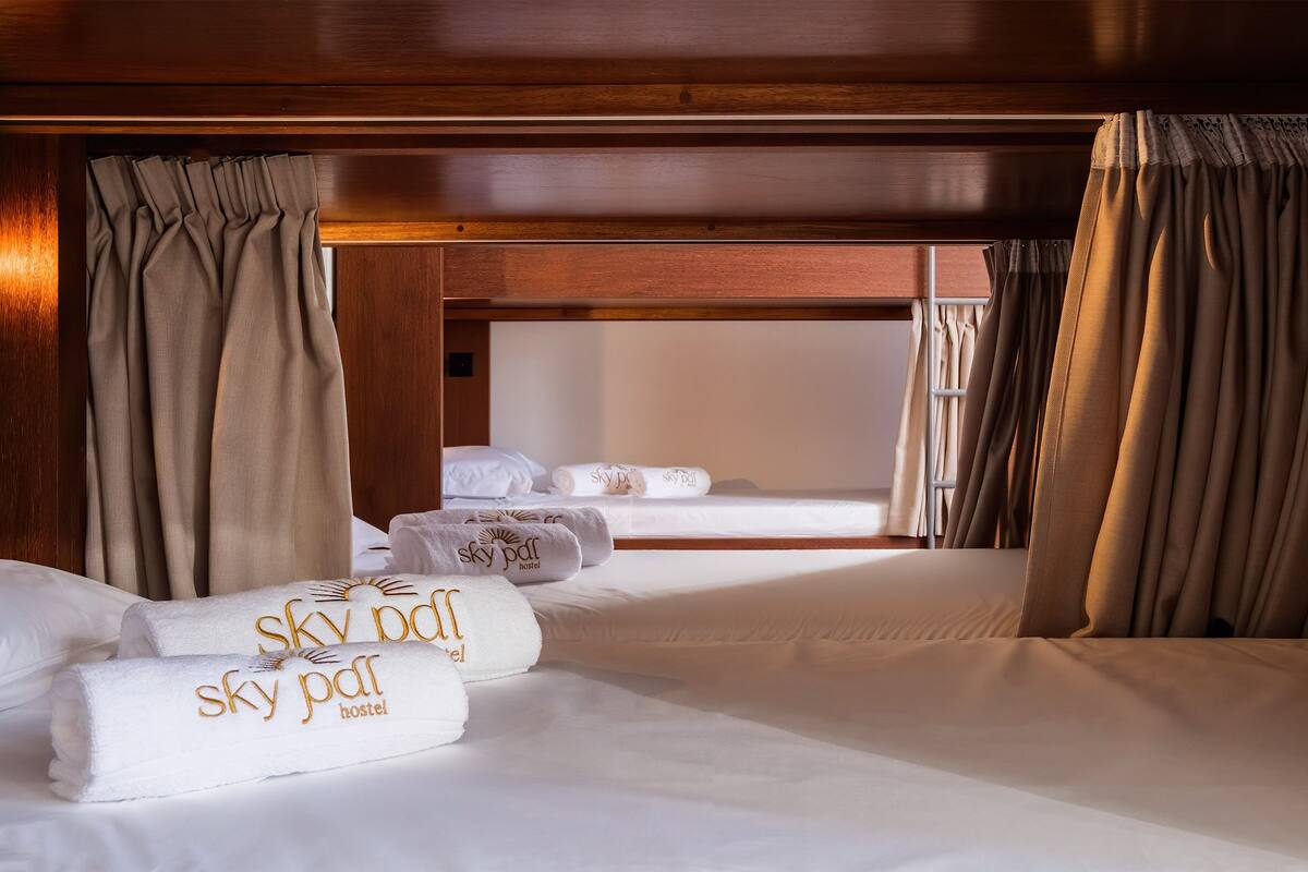 A bunk bed room with a wooden ceiling and beige curtains. There are towels with 'sky pdl hostel' embroidered on them.