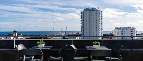 A terrace with four chairs and two potted plants, overlooking a construction site and a tall building by the sea.
