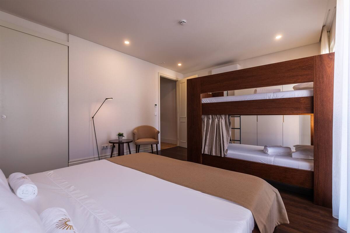 A hotel room with a large bed, a bunk bed, a chair, a small table, and a desk.
