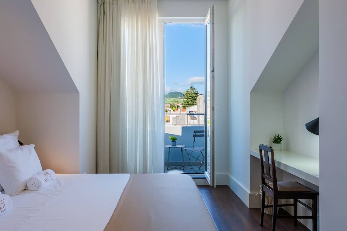 A hotel room with a large bed, a desk with a chair, a small table with a plant, and a balcony with a view of buildings and mountains.