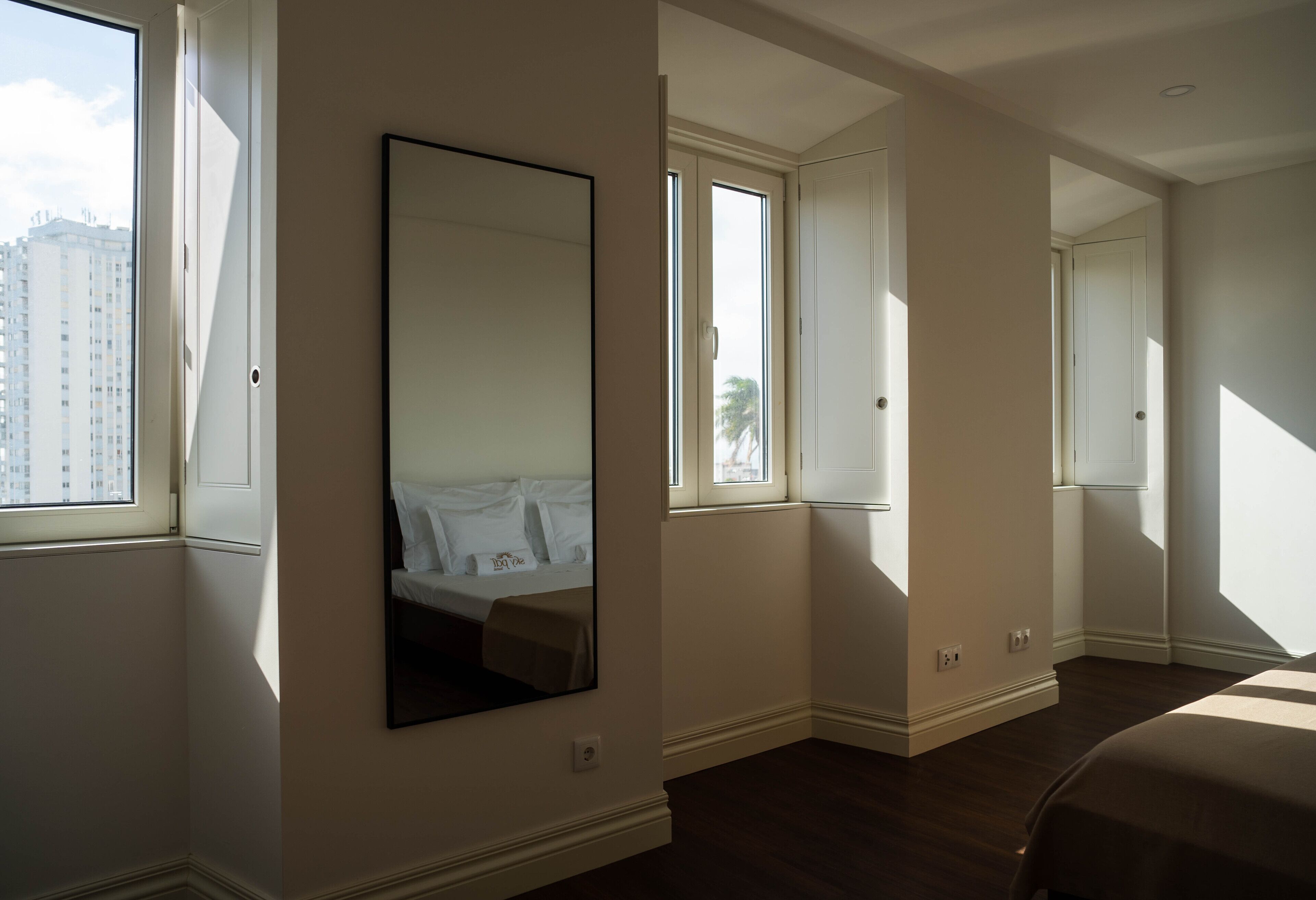 A hotel room with a large mirror, a bed, and multiple windows allowing natural light.