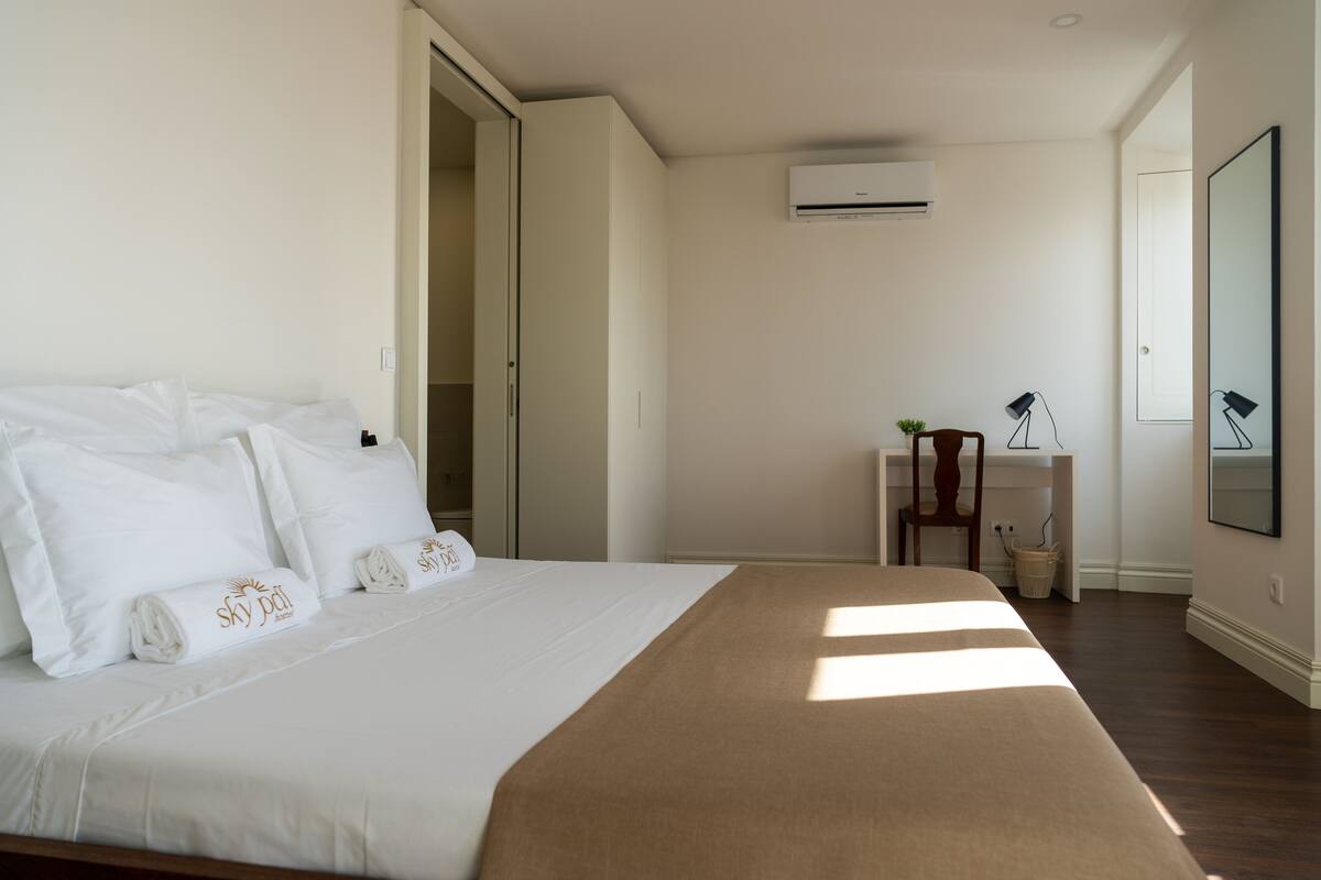 A bedroom with a bed, a desk, an air conditioner, and a mirror.