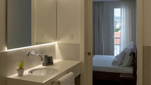 Een moderne badkamer met een grote spiegel, een wastafel en een handdoekhouder. Op het aanrecht staat een klein plantje en eronder een mand. Een deur leidt naar een slaapkamer met een bed en uitzicht op buiten.