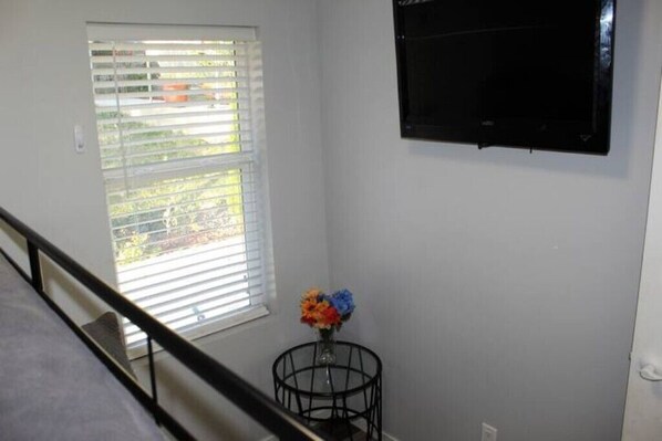 A room with a window, a black TV mounted on the wall, and a small table with a vase of flowers.