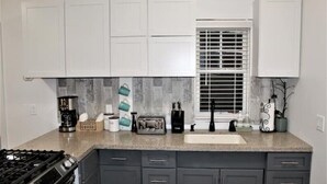 A kitchen with a gas stove, dark gray cabinets, a window with blinds, and various kitchen appliances and utensils on the countertop.