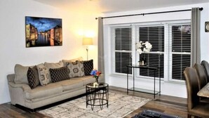 A living room with a sofa, a coffee table, a ceiling fan, a window with blinds, and a painting on the wall.