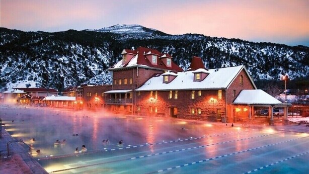 A hotel with a swimming pool, a large building with multiple windows, and a mountainous backdrop.