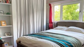 Ein Schlafzimmer mit Bett, Bücherregal, Fenster mit Vorhängen und Blick ins Grüne.