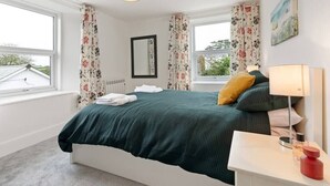 A bedroom with a bed, two windows, a bedside table with a lamp, and a painting on the wall. - Delightful Family Getaway Cornwall (Mawnan Smith)