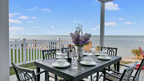 An outdoor dining area with a table set for four, overlooking a body of water, with a white fence and chairs. - Bayfront Home, Shared Pool/Spa, Gazebo, Playground (Port Isabel)