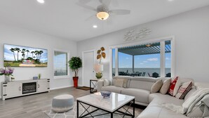 A spacious living room with a large window, a ceiling fan, and a view of the ocean. - Bayfront Home, Shared Pool/Spa, Gazebo, Playground (Port Isabel)