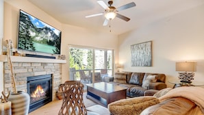 A living room with a fireplace, a television mounted above it, a ceiling fan, and a large window offering a view of the outdoors.