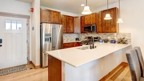 A modern kitchen with wooden cabinets, stainless steel appliances, a large island with a sink, pendant lighting, and a view of the living area.