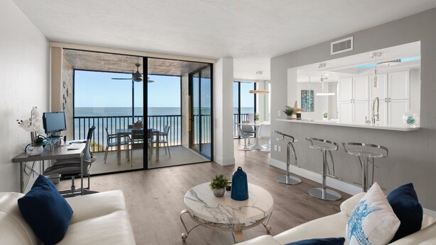 A modern living room with a large sliding glass door leading to a balcony with a view of the ocean. There is a dining area with a table and chairs, and a kitchen area with bar stools. - Get Ready for the Ultimate Beachfront Escape in Naples, Florida! (Naples)