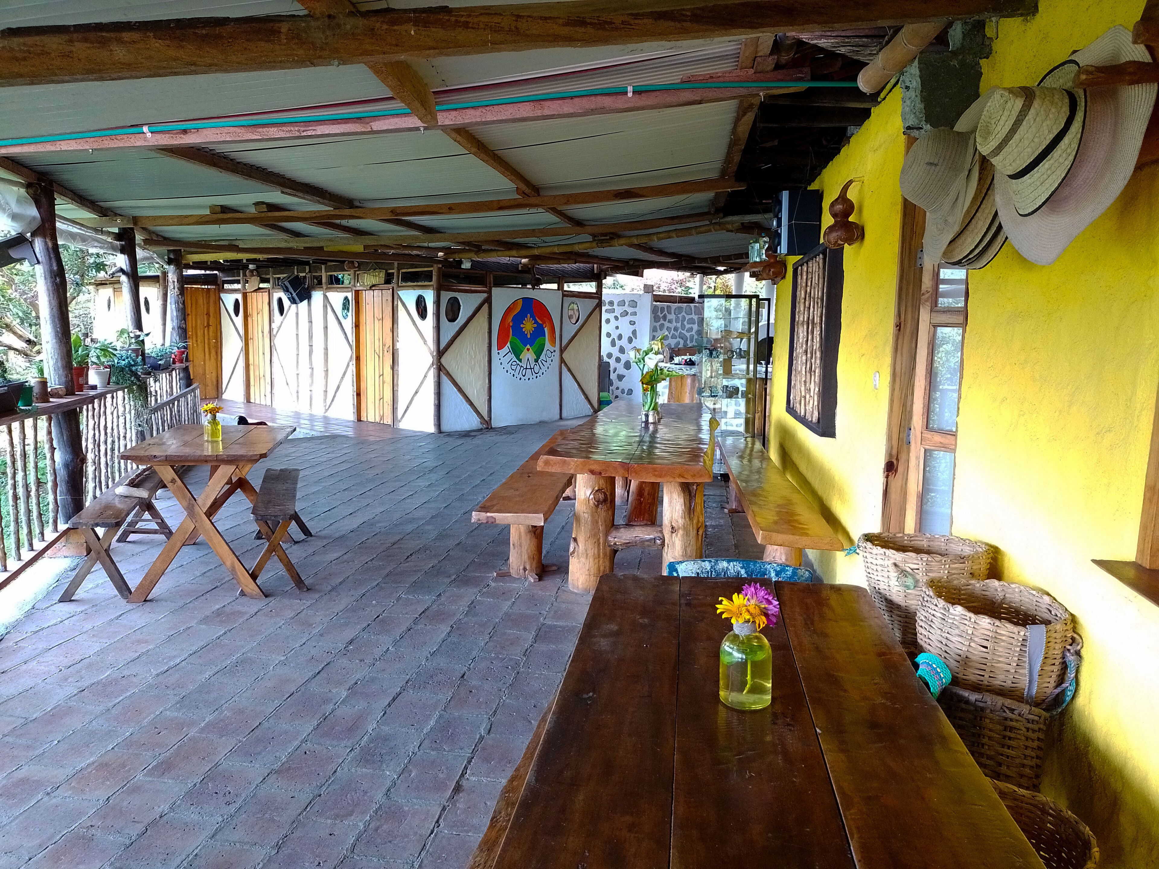 Una terraza exterior de madera con mesas y bancos, una pared amarilla y un mural.