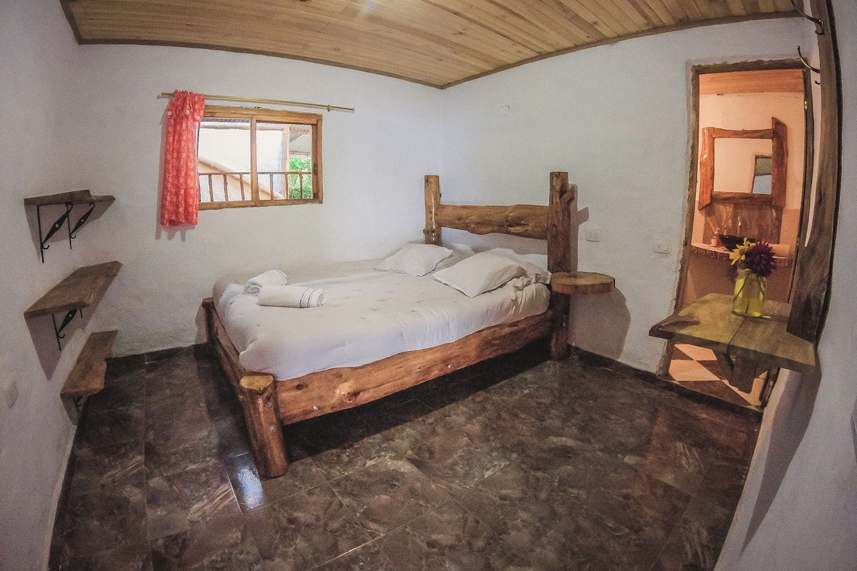 Un dormitorio rústico con una cama de madera, un ventanal con cortina roja y un banco de madera.