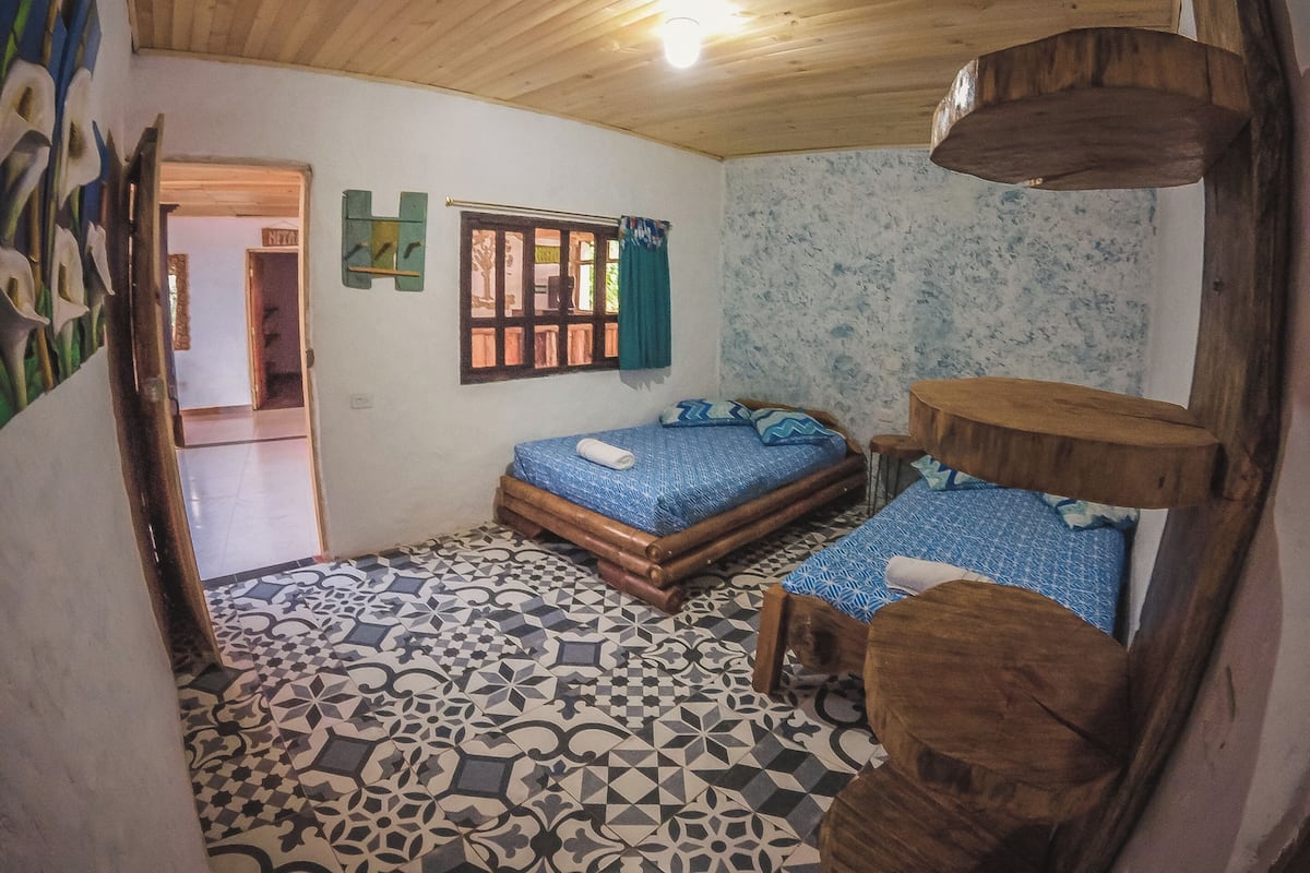 Una habitación pequeña y acogedora, con techo de madera y suelo de baldosas estampadas. Tiene una cama con ropa de cama azul y una pequeña mesa de madera.