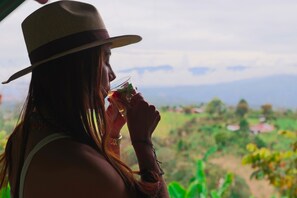 Une personne portant un chapeau et tenant un verre à la main, avec en arrière-plan un paysage verdoyant et des montagnes.
