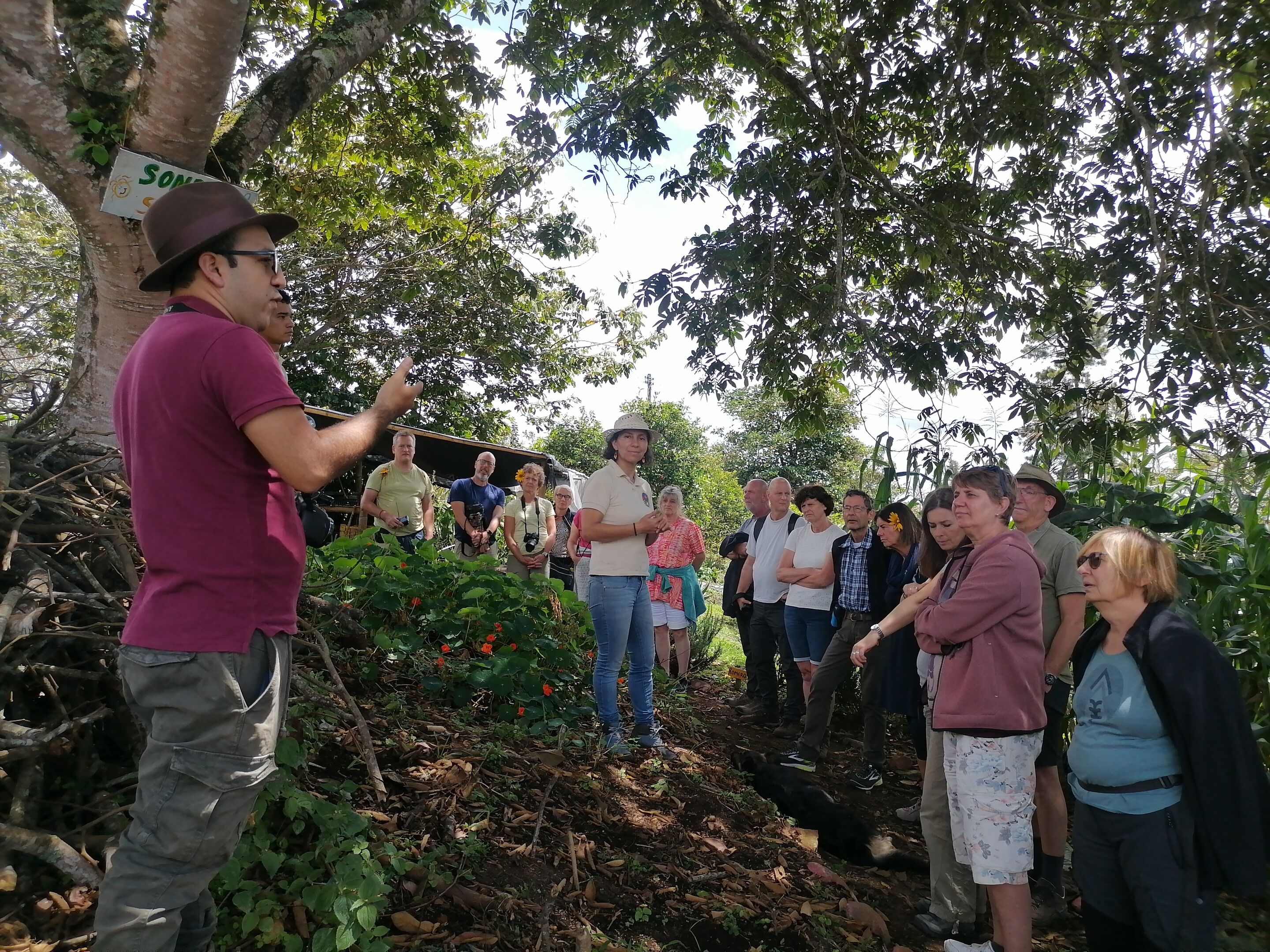 A group of people gathered outdoors, standing on a dirt path surrounded by trees and foliage.