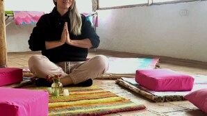 Una persona asseguda amb les cames encreuades sobre un estora de colors en una habitació amb bigues de fusta, una finestra i coixins roses.