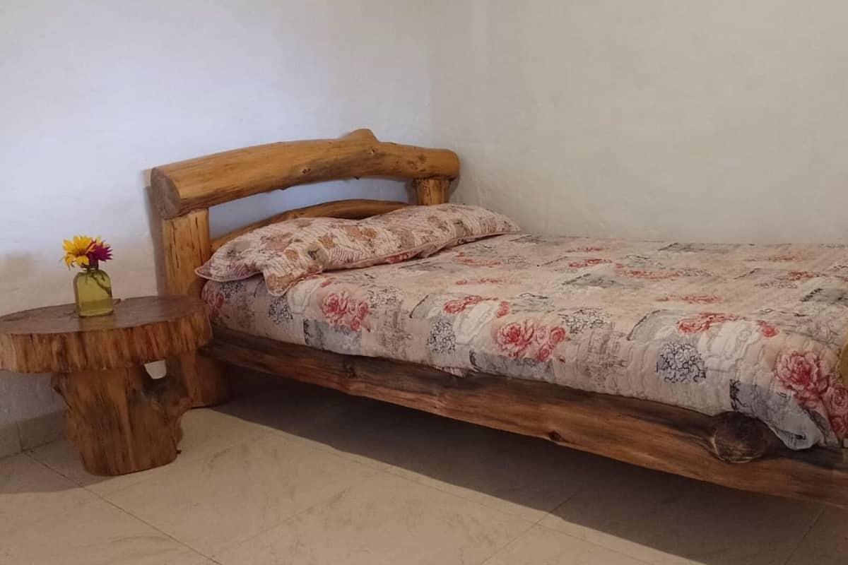 Una cama de madera con colcha estampada, una mesita de noche de madera con un florero y una pared lisa.
