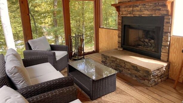 Una habitación acogedora con mobiliario de mimbre, chimenea de piedra y ventanas amplias con vistas a árboles.