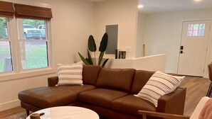 Una sala de estar con un sofá seccional marrón, una mesa de centro redonda y un sillón blanco. Hay un ventanal con persianas, una planta y una estantería con libros.