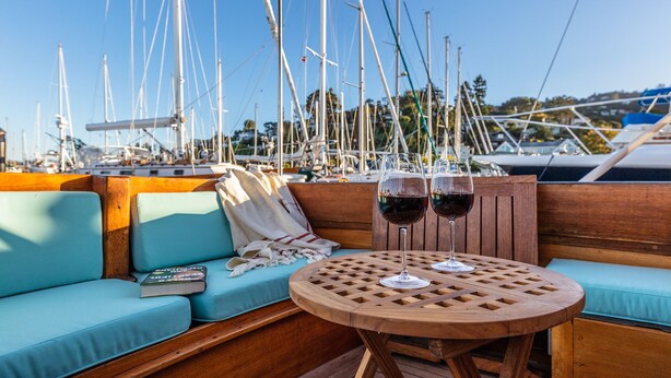 Une terrasse en bois avec une table ronde et un banc rembourré, deux verres de vin rouge et une télécommande posée sur le banc.