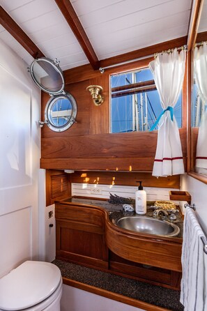 A compact, wooden cabin-style bathroom with a toilet, a sink with a vanity, a mirror, and a window with a view of the sea.