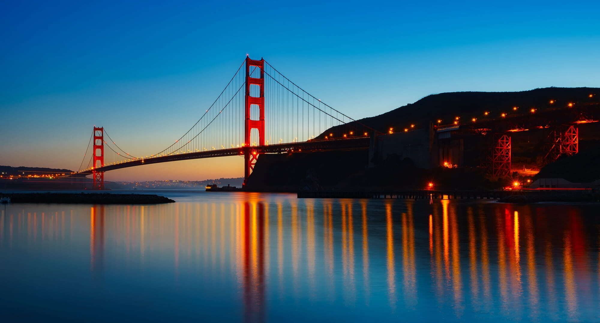 A bridge with a red structure spanning over a body of water, reflecting lights.