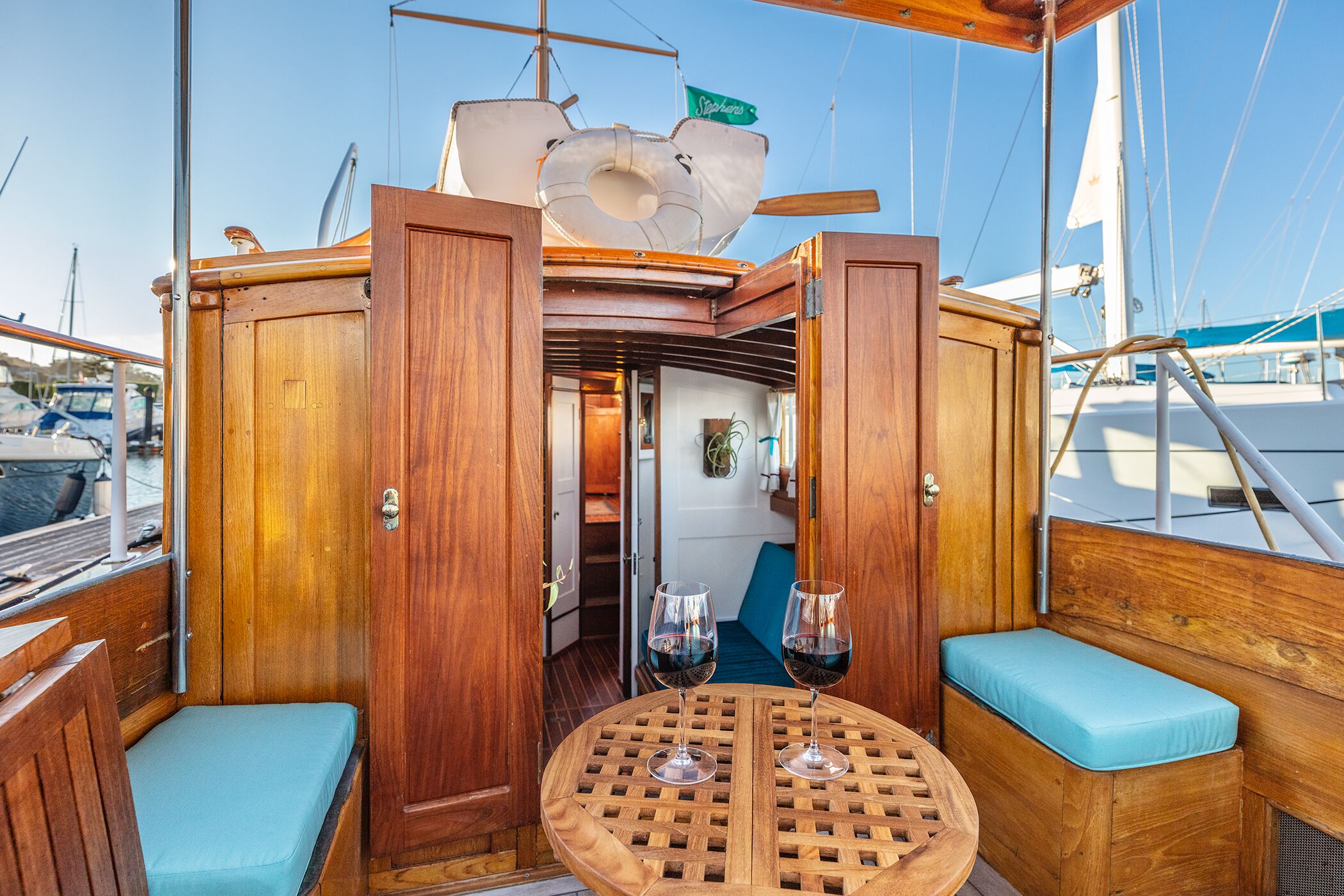 A wooden deck with a round table and two cushioned seating areas, a door leading to an interior space, and a sail visible in the background.