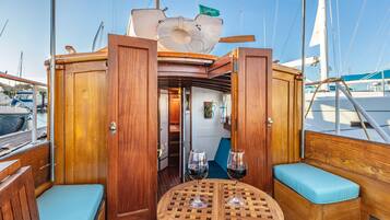A wooden deck with a round table and two cushioned seating areas, a door leading to an interior space, and a sail visible in the background.