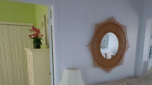 A living room with a sofa, a small wooden side table with a lamp, a mirror on the wall, and a door leading to another room. - Villa vanel (West End)