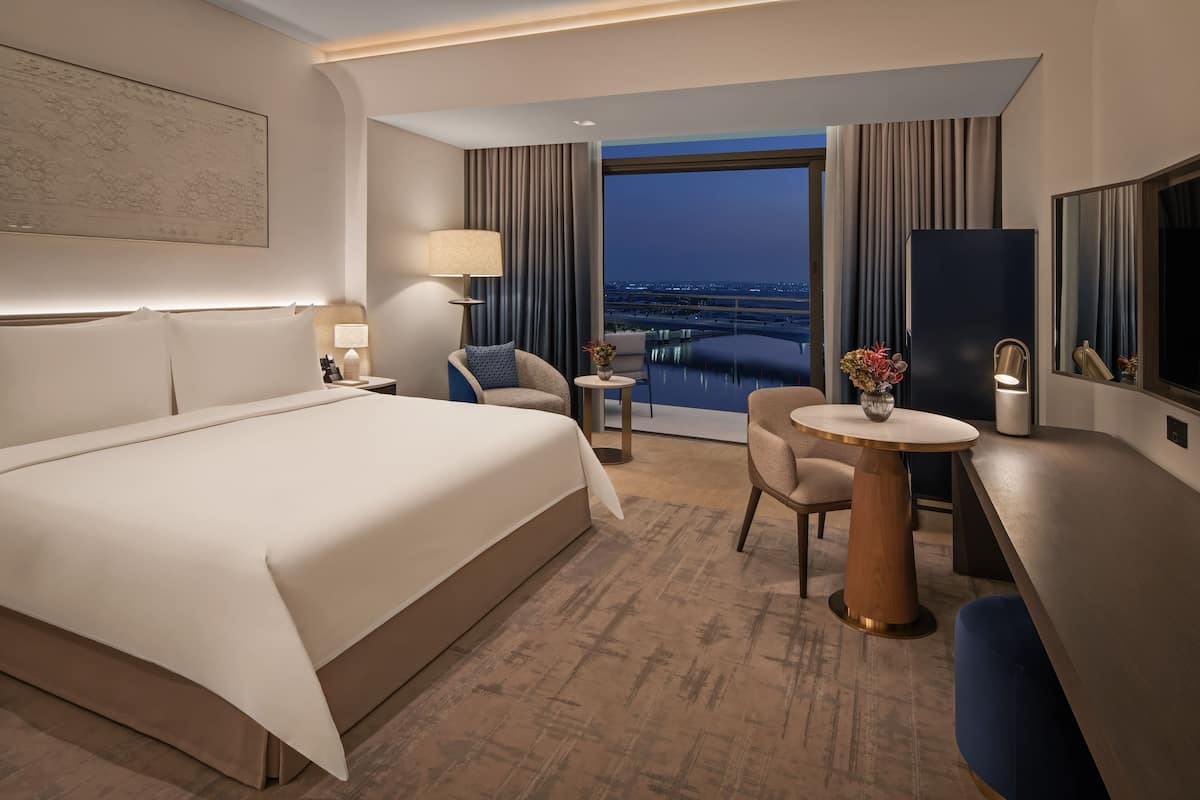 Een moderne hotelkamer met een groot bed, een bureau, een stoel, een kleine tafel met een vaas, en een groot raam met uitzicht op de stad.