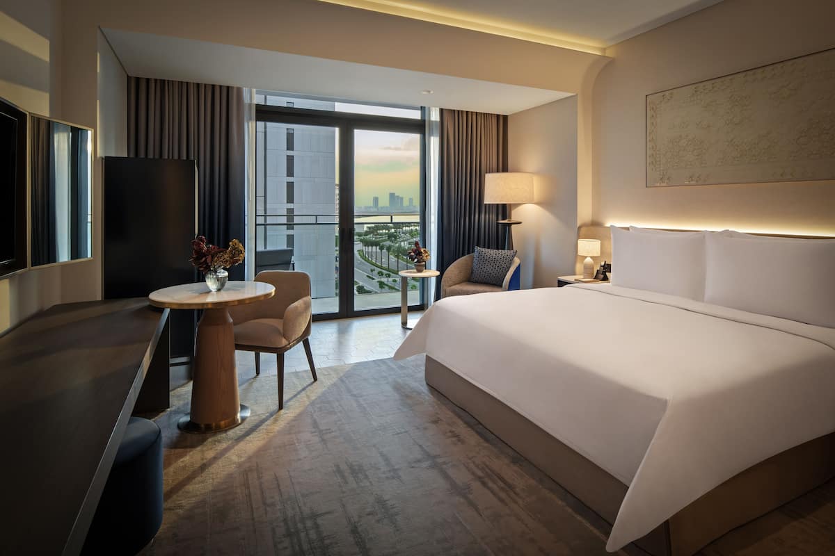 Een hotelkamer met een groot bed, een bureau, een stoel, een bijzettafeltje met een vaas, een lamp en uitzicht op de stad.