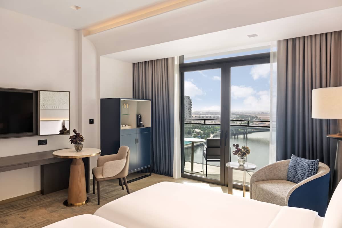 Een moderne hotelkamer met een groot bed, een bureau, een stoel, een klein tafeltje met een lamp, een televisie en uitzicht op een stadslandschap door grote ramen.