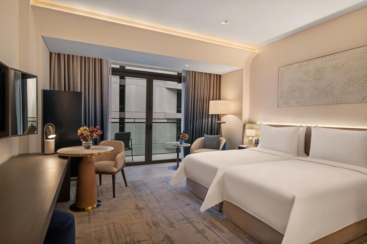 Een hotelkamer met een bed, een bureau, een stoel, een kleine tafel met bloemen, een lamp en een groot raam.