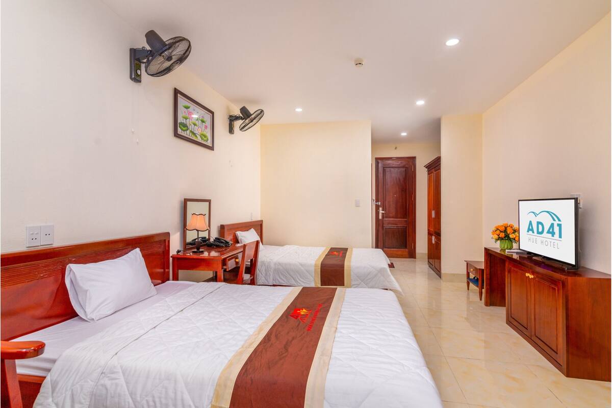 A hotel room with two beds, a desk, a chair, a TV, and a wall-mounted fan.
